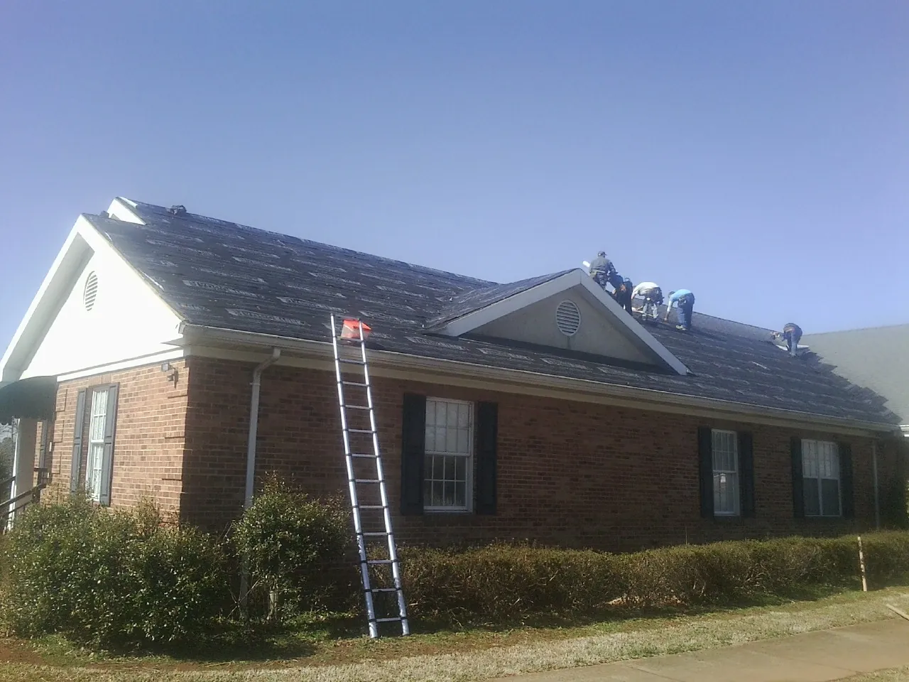 Crew tearing shingles off a brick ranch roof with a ladder set — Emergency Roof Repair work by our crew in Clarks Mills