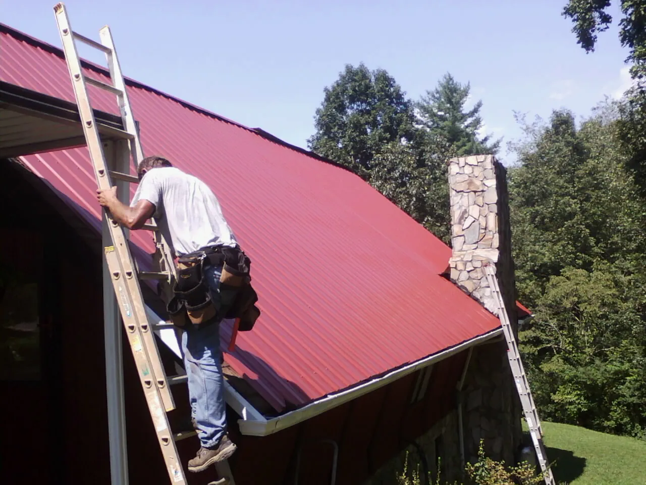 Roofer on a ladder beside a red metal roof and stone chimney — Soffit & Fascia Repair by Roofing Brothers in Clarks Mills
